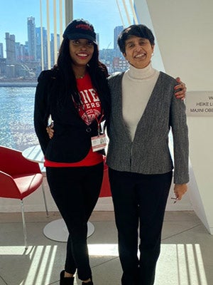 Two women standing shoulder to shoulder in front of a window with water and tall buildings in the background.