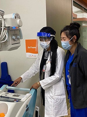 two women wearing masks looking down at images on an X-ray machine.