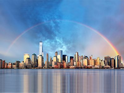 photo of New York City skyline with water in the foreground and a rainbow in the blue sky above the buildings