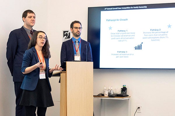 Two male and one female student dressed in business attire stand next to a podium and screen. The woman is gesturing and speaking.