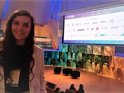 A picture of Ina Gjika at the opening plenary of the conference with the stage and big screen of sponsors behind her.
