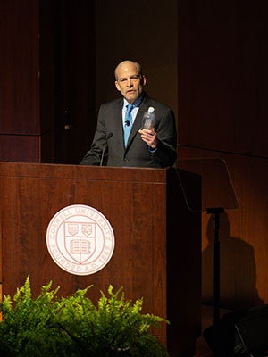 A man standing behind a podium, holding up a plastic bottle, and speaking.