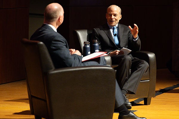 Two men on stage sitting in armchairs angled to face one another and the audience.