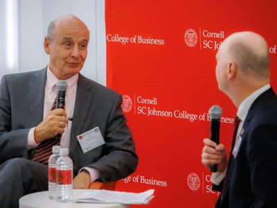 One man speaking into a microphone and looking at another man seated adjacent to him at a small table.