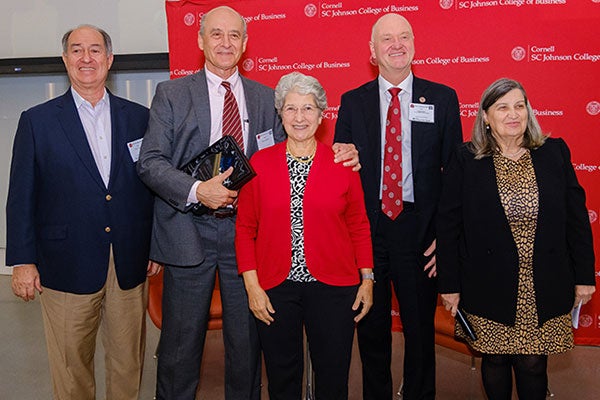 5 people standing and posing for a photo in front of a red backdrop with the Cornell University logo and the text: Cornell SJ Johnson College of Business.