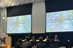 A conference room with panelists at a table and large presentation screens on the wall behind them.