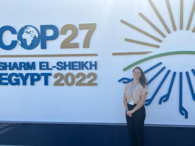 Courtney is standing to the left of a COP27 Entrance masthead which reads “COP27 Sharm El-Sheikh Egypt 2022”.