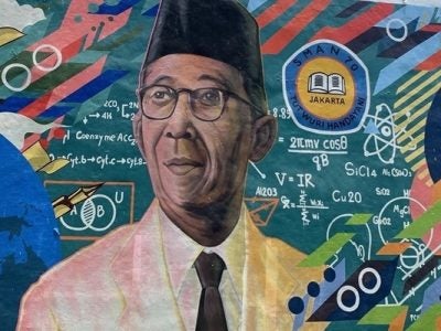 A mural featuring the head of an Indonesian man with glasses, wearing a traditional black peci cap, white jacket and black tie surrounded by colorful educational symbols from math and science.