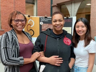 Molebi, JewleKrystal and Sheryl stand in front of a BaldGoddess trifold in Sage Hall's atrium.