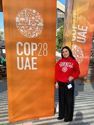 Rafaela Balde&oacute;n standing outside next to a large orange banner with the words: COP28 UAE.