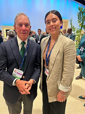 an older man and a young woman standing next to each other outside with many other people in the background.