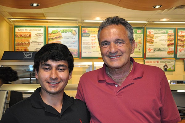 Upper body shot of two men standing side by side in front of a Subway counter.