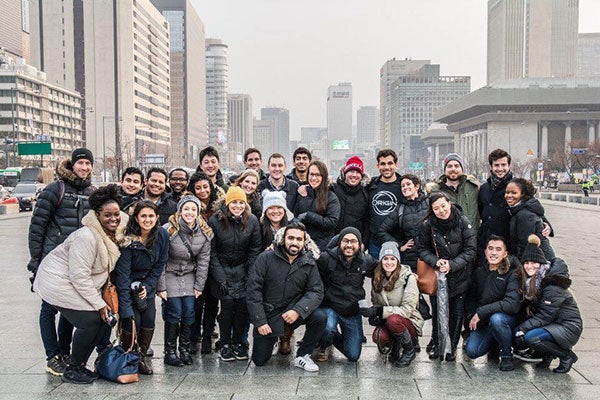 A large group of students bundled up in winter coats pose for a photo on a city square surrounded by tall buildings.