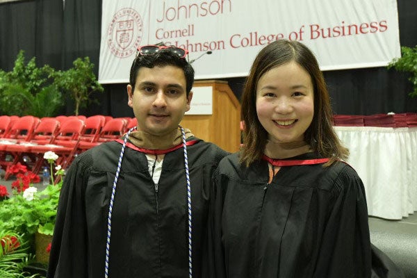 A man and a woman standing wearing graduation gowns with seating and flowers in the background below a banner with the text Johnson | Cornell | SC Johnson College of Business.