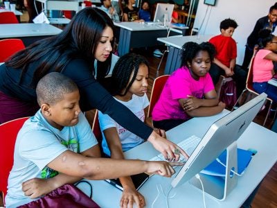 photo of Hilary Lashley Renison standing and leaning between middle-school-aged children who are sitting at a table and looking at a computer monitor, looking at and pointing to the screen