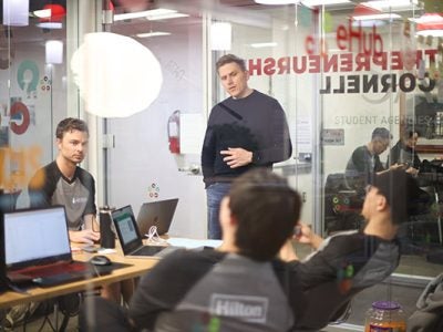 4 students at a table with laptops in a breakout room, one standing, three sitting.