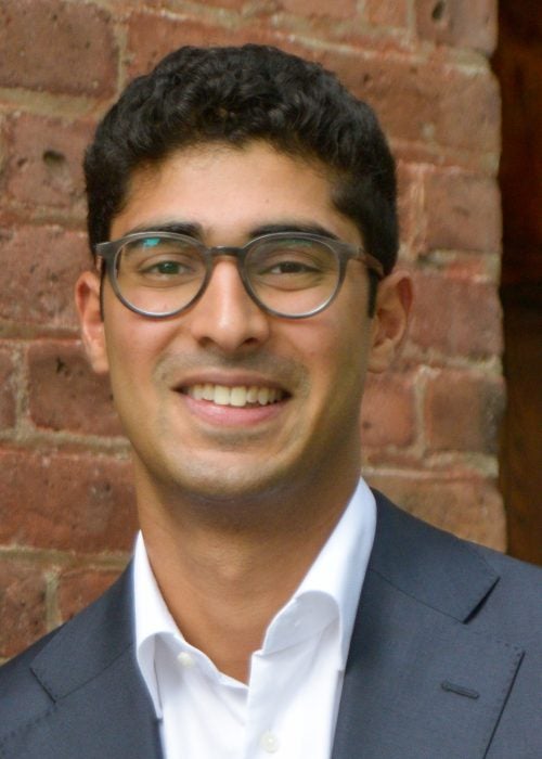 Roshen Sebastian, a young man in business attire against a brick backdrop, smiles as part of the Cornell MBA Class of 2024