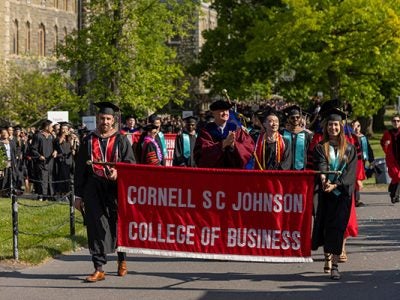 Students and faculty in caps and gowns holding and walking behind a Cornell SC Johnson College of Business banner.