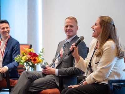 Two men and one woman seated on a stage. The woman is speaking into a microphone.