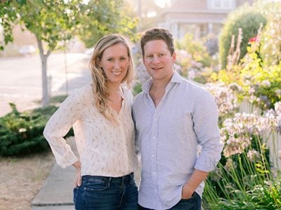 Julia and Alex Littauer stand outside on a city street with foliage in the background sunny, spring day.