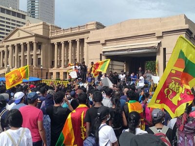 A large crowd of people, some waving Sri Lanka’s flag, facing government buildings and listening to speakers standing in front of those buildings.