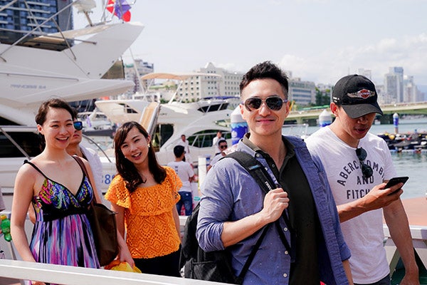 Steve Cheng in casual clothes with a backpack in foreground surrounded by several smiling young adults at a marina, boats in the background