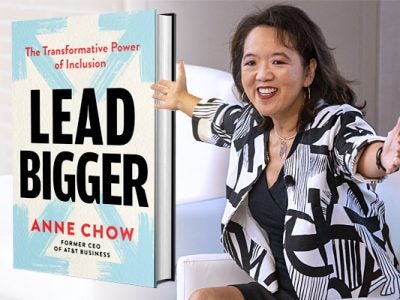 A woman in a black and white blazer with her hands outstretched to the right of an enlarged image of the book she authored.