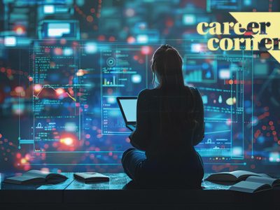 Silhouette from behind of a woman sitting on a table holding a laptop and looking at data and specifications on a wall-sized digital screen.