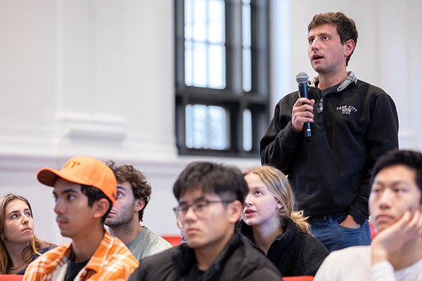 A male student standing at his seat in a lecture hall, surrounded seated students, speaks into a microphone.
