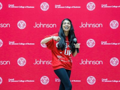 Trishala Dessai gesturing and smiling on stage wearing a red Gig-Up sweatshirt with a Johnson School banner in the background.