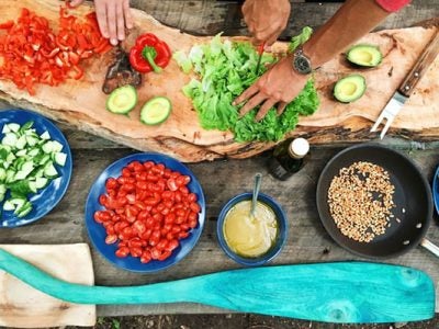 Components of a meal in preparation: cut up vegetables in bowls and on a cutting board along with a bowl of grains and of beans and a sauce. Also shows a person’s hands cutting up a green leafy vegetable