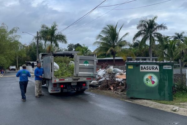 Two men load up a high-capacity truck with branches and tree bark. The truck is parked to the left on the right side of the road and there is a pile of bio-mass debris on the right side of the truck near a dumpster that reads &ldquo;Basura&rdquo;.