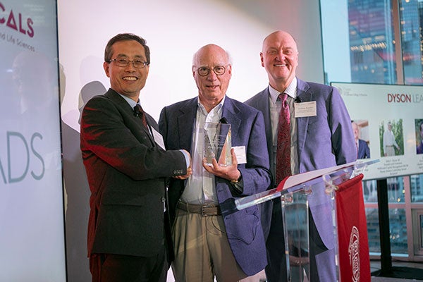 Three men standing side by side on a stage and smiling, with the one in the middle holding a crystal block with his name and the award name engraved on it (John S. Dyson &rsquo;65, Dyson Alumni Leadership Award).