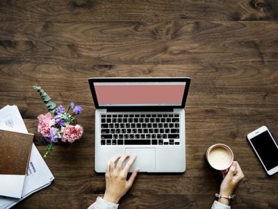 Photo from overhead of a woman on a laptop with a notebook, phone, and coffee on her desk