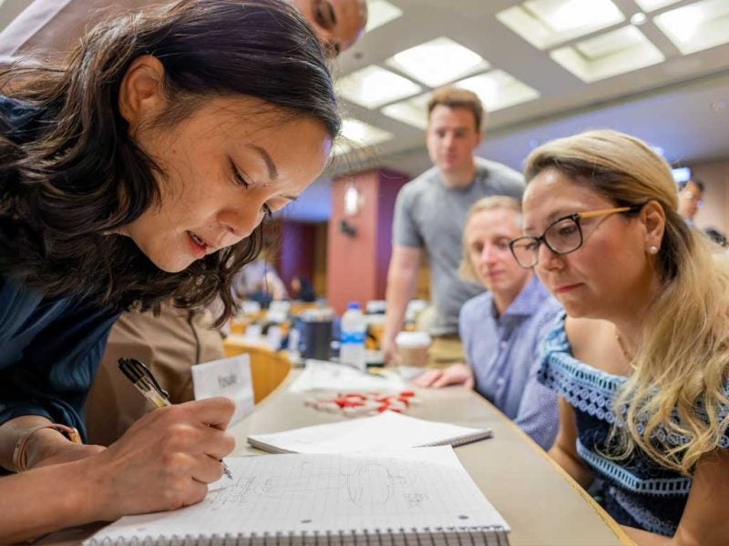 Women collaborate at a long table in a bright room, with the woman closest to the viewer writing in a notebook while others look on as part of the Cornell MBA curriculum