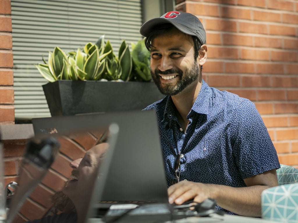 A young man in a button-up shirt and baseball hat smiles in a sunlit Dyson Atrium within Sage Hall as part of studying on his laptop for the Cornell MBA program