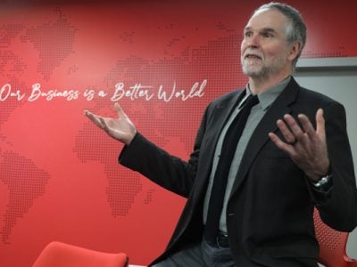 Professor John Doris sits on a stool with arms outstretched in front of a red wall with the text "Our Business is a Better World."