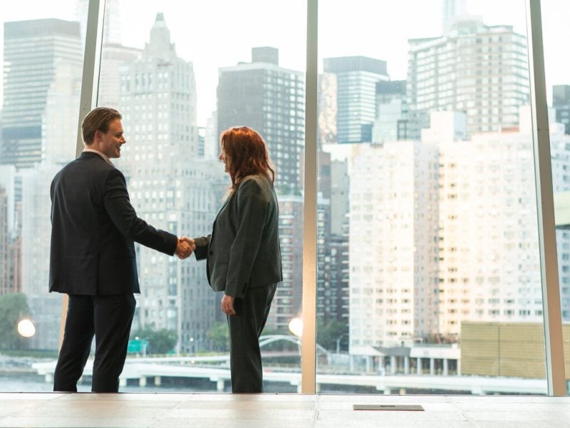 A man and woman dressed in business formal attire, enrolled in a Cornell Executive MBA program, shake hands in front of large glass walls showing the cityscape of New York City in the backgroun
