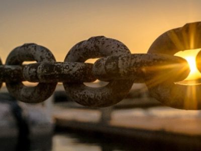 links of a large metal chain with the rising or setting sun shining through one of the links.