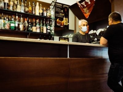 Photo of bartender wearing mask serves drink to hotel guest.