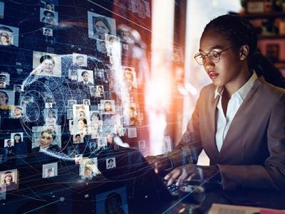 Business woman of color typing on a laptop with a hologram of a globe and connections to many headshots in the foreground
