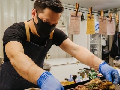 A chef working in a kitchen with a mask and gloves on, setting a plate of food onto a tray.