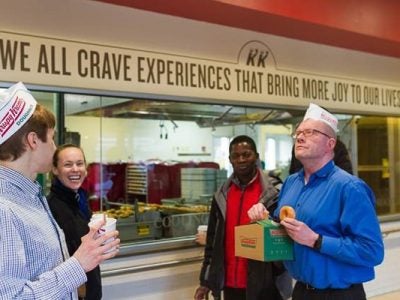 Photo of students and workers wearing Krispy Kreme hats in front of a sign that says "we all crave experiences that bring more joy to our lives"