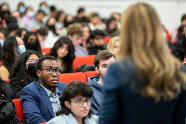 A young man in business casual attire listens attentively to a middle-aged woman professor in a mid-sized lecture hall as part of the Cornell MBA program