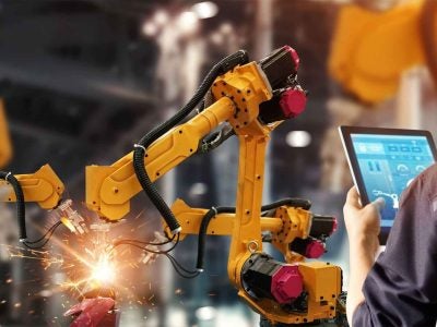 A worker holds a tablet as a robot arm welds.