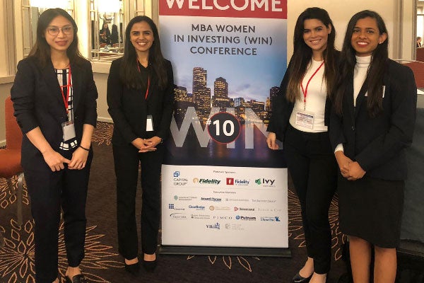 Four young women in business attire stand inside a hotel lobby alongside a banner welcoming visitors to the MBA Women in Investing (WIN) Conference as part of the Old Ezra Finance Club at Cornell