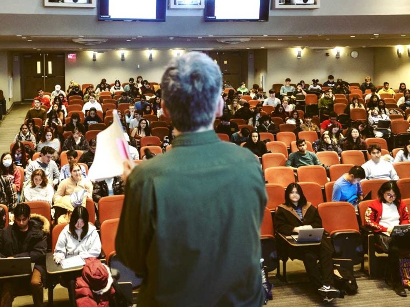 Audience-facing view of a professor teaching a class in a large lecture hall auditorium in Cornell's Statler Hall