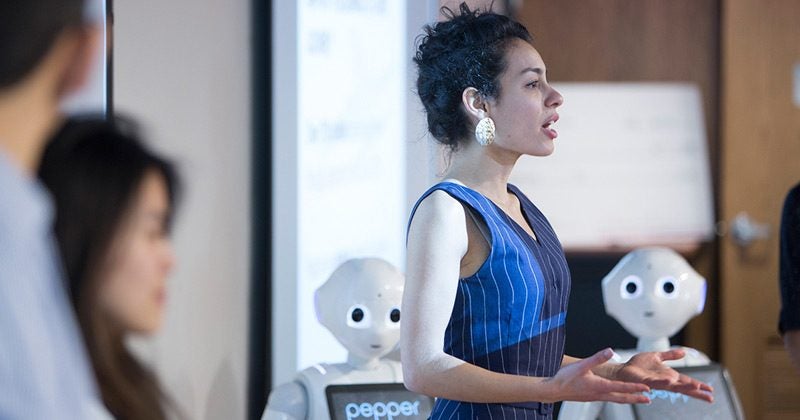 A woman speaks behind a podium near two Pepper robots