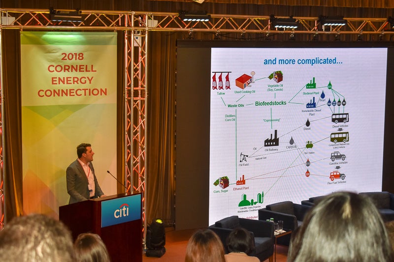 A man speaks at the podium with a detailed slide presenting a flowchart of processes