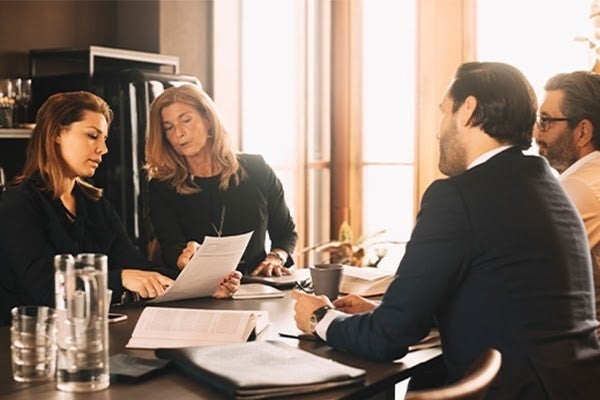 Two women and two men sit across from each other at a boardroom table and review documents.